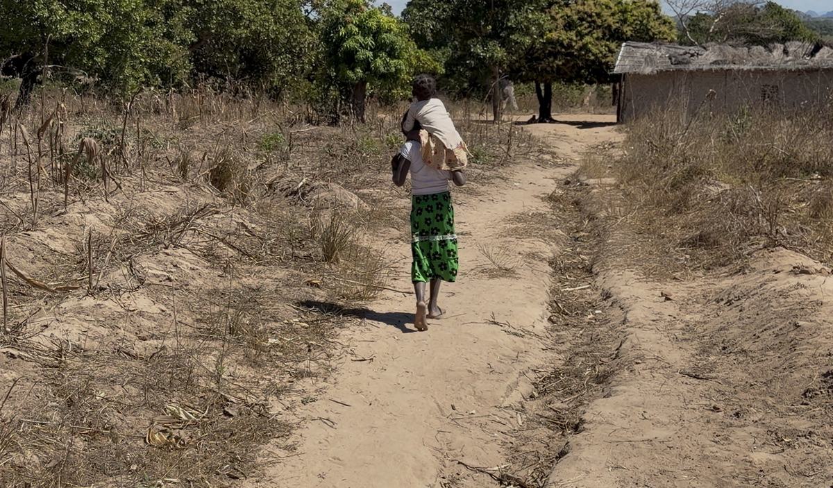 Mulher caminha carregando uma criança por um caminho de terra na aldeia