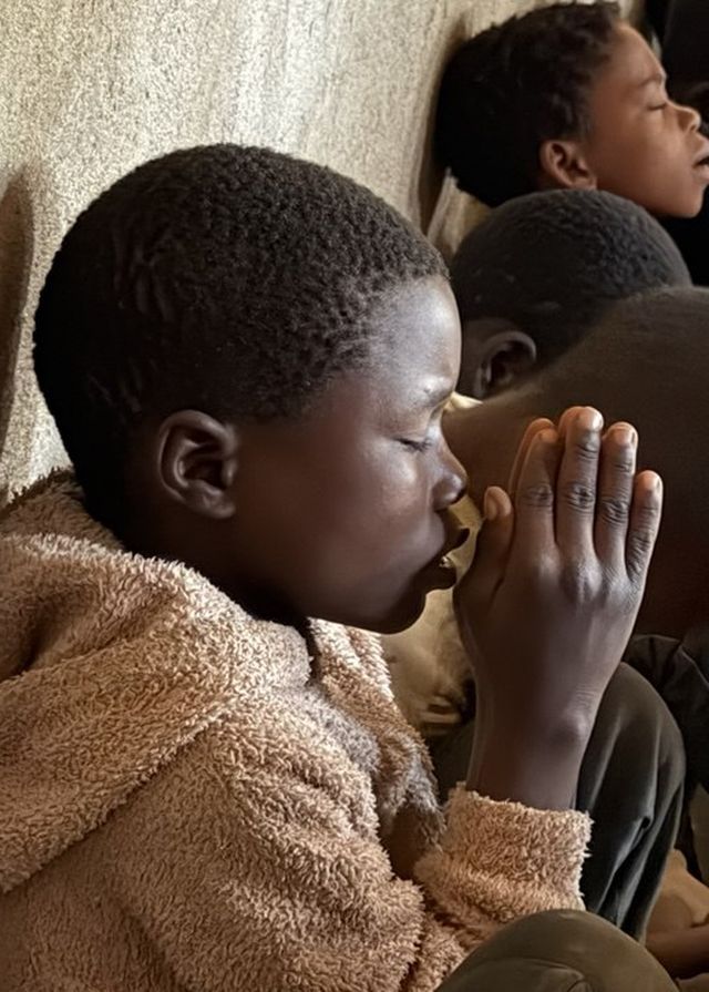 Menino de olhos fechados e mãos unidas em oração, entre outras crianças reunidas na aldeia de Maratane